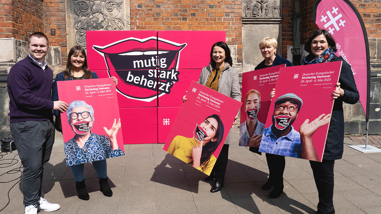 Plakate mit der Kampagne des Kirchentages werden von fünf lächelnden Menschen vor einer Kirche gehalten. Im Hintergrund eine große Fläche die ebenfalls das Motiv zeigt.