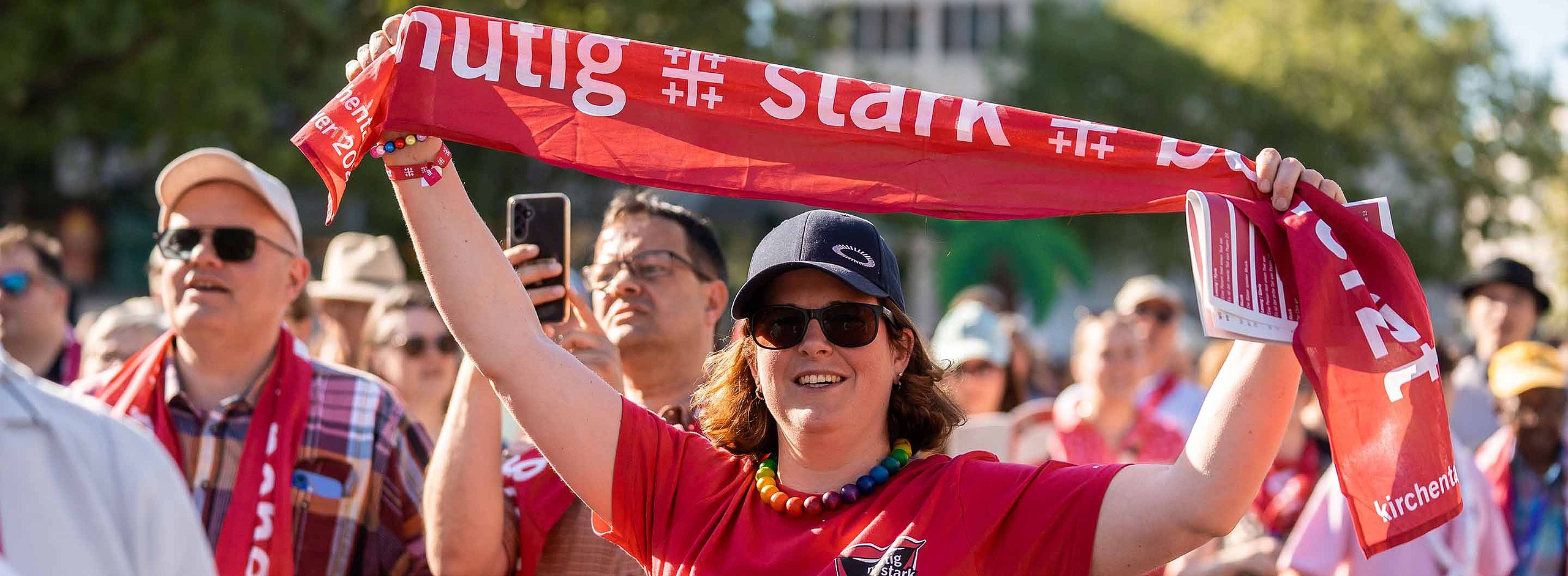 Teilnehmende beim Kirchentag in Hannover (Foto: Kirchentag/Weise)  Eine Frau mit Mütze, Sonnenbrille und rotem Kirchentagsshirt steht in der Menschenmenge und hält einen roten Kirchentagsschal in die Luft.