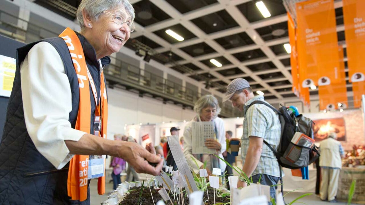 Alte Dame mit Kirchentagsschal lacht, vor ihr steht ein Blumenbeet mit kleinen Papierschildern, im Hintergrund das Marktgeschehen in einer Messehalle